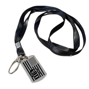 May include: A black lanyard with a silver metal clasp and a rectangular dog tag. The dog tag features an American flag design and the number "911" with a firefighter's helmet.