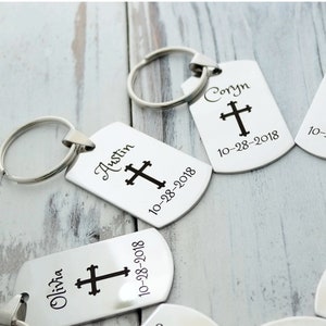 May include: Silver rectangular keychains with a cross design and personalized text. Each keychain has a name and date engraved, attached to a silver keyring. The keychains are displayed on a light-colored wooden surface.