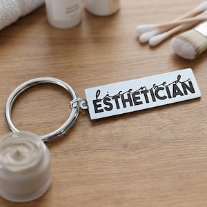May include: A silver-toned rectangular keychain featuring a split ring and a metal tag. The tag displays the text "licensed ESTHETICIAN" in a combination of black cursive and bold sans-serif lettering, centered on the smooth metallic surface.