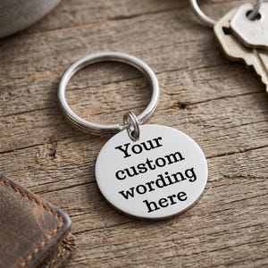 May include: A silver-colored round keychain with the text "Your custom wording here" in black. The keychain is attached to a silver ring and rests on a wooden surface. Keys are visible in the background.