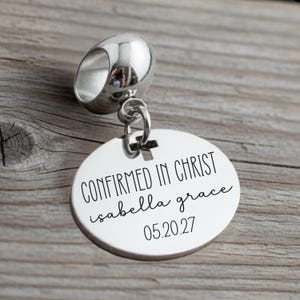 May include: A silver charm with a round pendant. The pendant has the words "CONFIRMED IN CHRIST" and the name "isabella grace" with the date "05.20.27" engraved in black. The charm has a cross design.