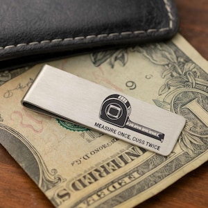 May include: A silver money clip with a black tape measure graphic and the text "MEASURE ONCE, CUSS TWICE." The clip rests on a dollar bill, with a black leather wallet in the background.