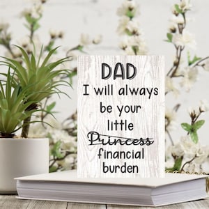 May include: White wooden sign with black text that reads "DAD I will always be your little Princess financial burden" with the word "Princess" crossed out.