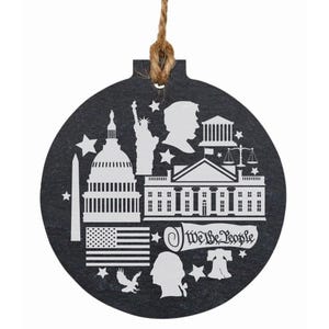 May include: A round, dark gray ornament with a white design featuring the U.S. Capitol, the Statue of Liberty, the White House, an American flag, and the text "We the People." A twine loop is attached for hanging.