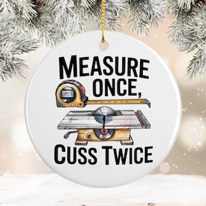 May include: White ceramic ornament with a gold string for hanging. The ornament features a tape measure and a table saw illustration with the text "MEASURE ONCE, CUSS TWICE" in black. A humorous gift for a carpenter or woodworker.