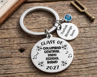 May include: A silver-toned metal keychain featuring a large circular charm engraved with "CLASS OF COLUMBIA CENTRAL HIGH SCHOOL BAND 2027" surrounded by musical notes and stars, a smaller circular charm with "Custom Message Here", and a blue crystal.