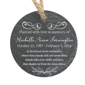May include: A round, dark gray slate ornament with a white inscription that reads "Planted with love in memory of Michelle Anna Farmington October 25, 1987 - February 3, 2024 in the forest of our memories, where time stands still and never flees. stands tall a tree of life and love, that shades us from the skies above."