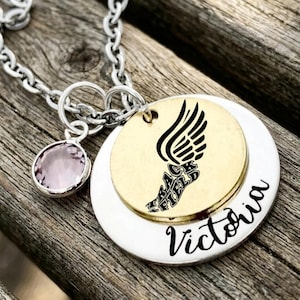 May include: A silver necklace with a round pendant featuring a gold disc with a black winged foot design and the words "Track Field". A smaller silver disc and a pink gemstone charm are also attached. The name "Victoria" is written in cursive.