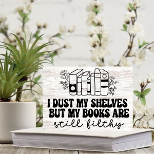 May include: A white wooden sign with the text "I DUST MY SHELVES BUT MY BOOKS ARE still filthy" in black. The sign features a line drawing of books and floral accents. The sign is resting on a white book.