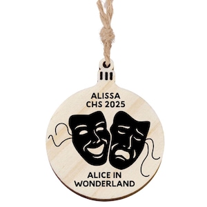 May include: A round wooden Christmas ornament with a natural wood finish. The ornament features black theater masks and the text "ALISSA CHS 2025" and "ALICE IN WONDERLAND". A natural twine loop is attached for hanging.