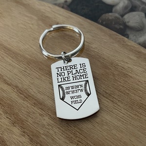 May include: A silver-colored keychain with a rectangular tag. The tag has the text "THERE IS NO PLACE LIKE HOME" above a baseball graphic and coordinates. Below the graphic is "WCHS FIELD". The keychain is on a wooden surface.