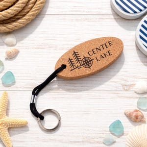 May include: Oval cork keychain with the text "CENTER LAKE" and a tree and compass design. The keychain has a black cord and a silver metal ring. The background includes seashells, sea glass, and rope.