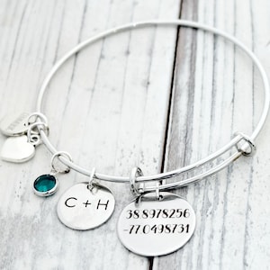 May include: Silver bangle bracelet with two charms. One charm has the initials "C+H" engraved on it. The other charm has the coordinates "38.8978256 -77.0498731" engraved on it. The bracelet also has a small teal blue gemstone charm and two silver heart charms.
