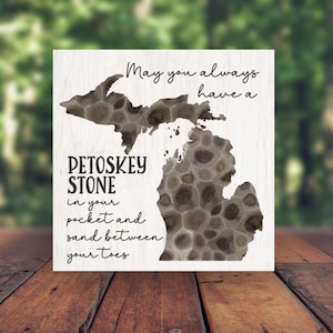 May include: A white wooden sign with a silhouette of the state of Michigan. The sign features the text "May you always have a Petoskey Stone in your pocket and sand between your toes."