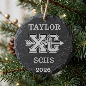May include: A round, dark gray slate ornament with a rough, natural edge. It features the engraved text TAYLOR, the letters XC with a winged foot icon, the text SCHS, and the year 2026. A brown twine loop is attached to the top for hanging.
