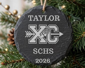 Könnte beinhalten: Ein rundes, dunkelgraues Schieferornament mit rauer, natürlicher Kante. Die Oberfläche ist mit dem Namen TAYLOR, den Buchstaben XC mit einem geflügelten Fuß-Symbol, dem Akronym SCHS und dem Jahr 2026 graviert. Eine Juteschnur ist oben befestigt.