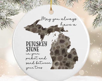 May include: Round white ceramic ornament with a gold hanging string. The ornament displays a map of Michigan filled with a Petoskey stone design. Text reads "May you always have a Petoskey Stone in your pocket and sand between your toes."