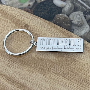 May include: A silver keychain with a rectangular tag. The tag has the text "MY FINAL WORDS WILL BE are you fucking kidding me?" in black. The keychain is on a wooden surface.