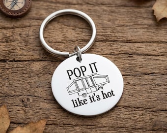 May include: A circular silver-toned metal keyring featuring a black engraved graphic of a pop-up camper trailer. The text "POP IT" is positioned above the camper illustration, and the phrase "like it's hot" is engraved below the camper.