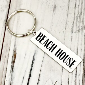 May include: A silver rectangular keychain with the words "BEACH HOUSE" in black, attached to a silver key ring. The keychain is angled on a light wood-grain surface.