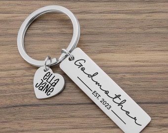 May include: A silver-toned metal keychain featuring a circular ring, a heart-shaped charm engraved with the name Ella Jane, and a rectangular tag engraved with the text Godmother EST. 2023 in a black script font.
