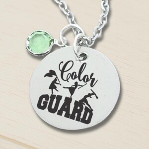 May include: Silver-toned necklace with a round pendant featuring the words "Color Guard" and silhouettes of performers. A small green gemstone charm hangs near the pendant. The chain is a classic link style.