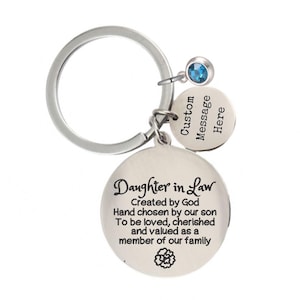 May include: A silver-colored keychain with a large circular pendant engraved with the words "Daughter in Law." It includes a smaller pendant with the words "Custom Message Here" and a blue gemstone. The keychain is designed to be a sentimental gift.