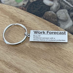 May include: A silver metal keychain with a circular ring and a rectangular tag. The tag reads "Work Forecast" with the text "Busy AF, Scattered sarcasm with a strong chance of muttered profanities."