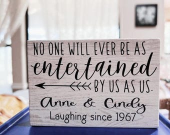 May include: A rectangular wooden sign with the text "NO ONE WILL EVER BE AS entertained BY US AS US. Anne & Cindy Laughing since 1967" in black lettering. The sign has a light grey, weathered appearance.