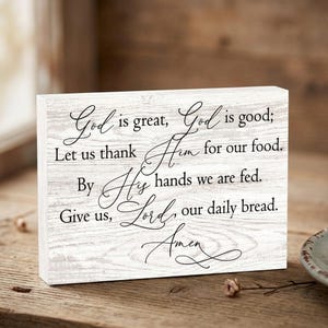 May include: A rectangular wooden sign with a white-washed finish. The sign features elegant black script that reads "God is great, God is good; Let us thank Him for our food. By His hands we are fed. Give us, Lord, our daily bread. Amen."