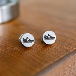 May include: A pair of silver stud earrings with a running shoe design. Each earring features a white circular face with a black running shoe silhouette and a heart. The earrings are displayed on a wooden surface.