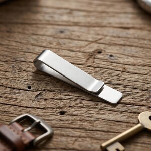 May include: A brushed silver-tone metal money clip with a simple, elongated, folded design. The clip features a smooth, matte finish and a clean, minimalist rectangular shape with rounded edges, designed to hold currency or cards securely.