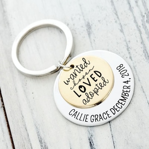 May include: A silver-toned metal keychain featuring two circular discs. The smaller gold-toned disc is engraved with the words wanted, chosen, LOVED, and adopted. The larger silver disc is engraved with CALLIE GRACE DECEMBER 4, 2018.