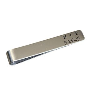 A silver metal tie clip with a personalized inscription on it.