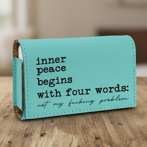 May include: A rectangular turquoise leather case with black text that reads: inner peace begins with four words: not my fucking problem. The case features visible stitching along the edges and a side flap, presenting a smooth, matte finish.
