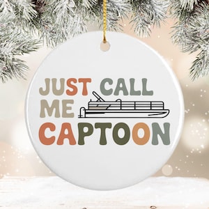 May include: A white ceramic ornament with the text "JUST CALL ME CAPTOON" in shades of brown, green, and orange. A black and white illustration of a pontoon boat is in the center. A gold string is attached for hanging.