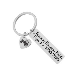 May include: Silver keychain with a heart-shaped charm featuring a tractor. The charm is attached to a rectangular tag with the text "Farming Heavens Fields Papa Joe. 1935-2023".