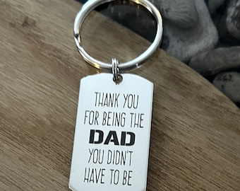 May include: A silver-colored dog tag keychain with the words "THANK YOU FOR BEING THE DAD YOU DIDN'T HAVE TO BE" in black and white. The keychain has a silver ring and is displayed on a wooden surface.