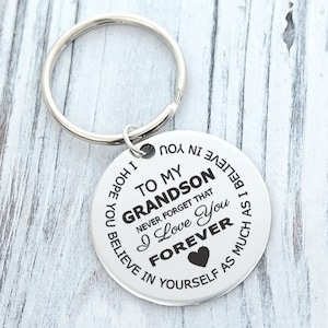 May include: A silver-colored keychain with a round pendant. The pendant has the text "TO MY GRANDSON, I Love You FOREVER" and other phrases. A heart symbol is also present. The keychain is on a white wooden surface.