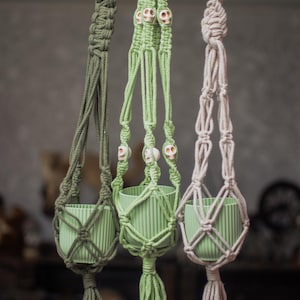 May include: Three macrame plant hangers in shades of green and gray, suspended from a wooden rod. Each hanger holds a ribbed, light green pot. The central hanger features skull beads, adding a unique touch to the decor.