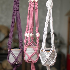 May include: Three macrame plant hangers in purple, pink, and white, each holding a ceramic pot. The pink hanger features skull beads. The hangers are suspended from a wooden dowel, showcasing a bohemian style.