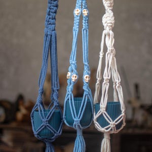 May include: Three macrame plant hangers suspended from a wooden branch. The hangers are made of dark blue, light blue, and off-white cord. Each hanger holds a teal ribbed plastic pot. The light blue hanger features small white skull beads.