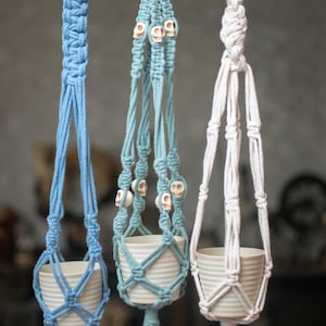 May include: Three macrame plant hangers suspended from a wooden dowel. The hangers are made of blue, teal, and white cord. The teal hanger features small white skull beads woven into the design. Each hanger holds a small, ribbed white ceramic pot.