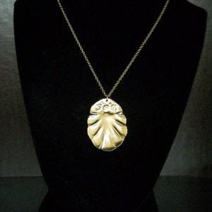 May include: A gold necklace with a large, ornate pendant shaped like a seashell. The pendant has a textured surface and a raised design.