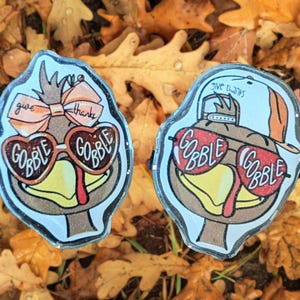 May include: Two Thanksgiving-themed turkey pins. One turkey wears a bow and heart-shaped glasses that say "GOBBLE GOBBLE." The other turkey wears a hat and sunglasses that say "GOBBLE GOBBLE." Both pins have the words "give thanks."