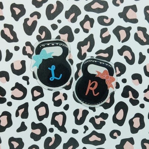 May include: Two black kettlebell-shaped keychains with a blue and red bow on each. The keychains are on a white background with a leopard print pattern. The keychains have the letters "L" and "R" on them.