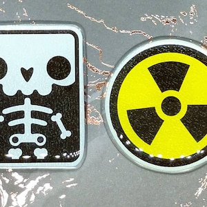 May include: Two metal pins with a spooky theme. One is rectangular with a skeleton x-ray design in white on a black background. The other is round with a yellow radiation symbol on a black background.