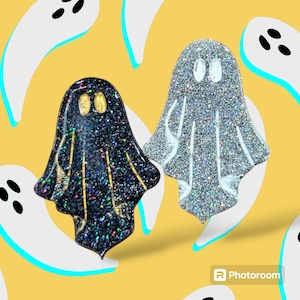 May include: Two glitter ghost pins, one black with gold accents and one silver. The pins are on a yellow background with white pear-shaped outlines.