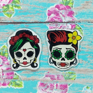 May include: Two colorful sugar skull stickers with black hair, green eyes, and red lips. The stickers are on a blue and white wooden background.
