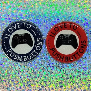 May include: Two round buttons with a black and white video game controller graphic. The text "I LOVE TO PUSH BUTTONS" is written around the controller on both buttons. One button is blue and the other is red.